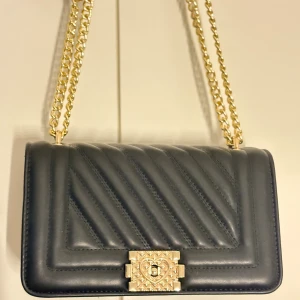 Svart quiltad axelväska med guldkedja - Snygg svart axelväska i quiltat skinn med guldfärgad kedja och dekorativt lås framtill med hjärtdetalj. Väskan har en stilren, boxig form och passar perfekt när du vill lyfta din outfit med något extra. Perfekt storlek för det viktigaste. Aldrig använt.