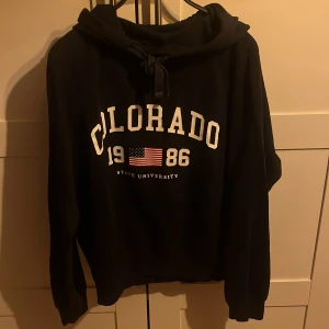 Mörk blå Colorado Gina Tricot S - Svart hoodie från Gina Tricot i storlek S med tryck 'Colorado 1986 State University' och amerikansk flagga framtill. Tröjan har huva med snörning och långa ärmar. Perfekt för chill dagar och enkel att matcha med jeans eller leggings.