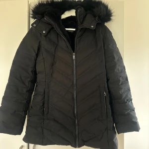 Svart pufferjacka med fuskpäls från Zara - Säljer en svart pufferjacka från Zara i storlek L. Den sitter definitivt mer som M vill jag säga! Jackan har huva med fuskpäls, dragkedja framtill och mysigt foder. Perfekt för kalla dagar med sin långa modell och quiltade design. Snygg och stilren look som håller dig varm hela vintern.