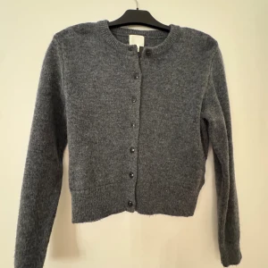 Grå kofta från H&M i ull XS - Mysig grå kofta från H&M i storlek XS, tillverkad i mjuk ullblandning. Modellen är croppad med rund hals och knappar framtill. Perfekt att slänga över en t-shirt eller linne när det är kyligt ute. Klassisk och enkel design som funkar till allt.