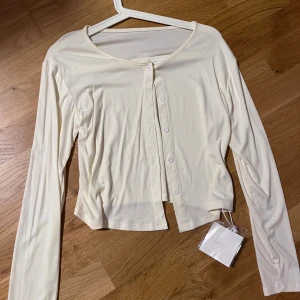 Krämvit / krämgul kofta från LALA - Säljer en krämvit/ krämgul långärmad topp från LALA med rund halsringning och knappar framtill. Toppen är croppad och har en mjuk, tunn känsla i materialet. Perfekt att styla med jeans eller kjol för en enkel och fräsch look. HELT NY MED PRISLAPP