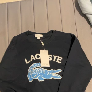Mörkblå sweatshirt från Lacoste - Snygg mörkblå sweatshirt från Lacoste med stort vitt tryck och en blå krokodil på bröstet. Tröjan har rund hals, långa ärmar och ribbade muddar. Regular fit och klassisk stil, perfekt för dig som gillar streetwear och ikoniska märken.