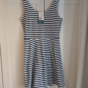 Randig kortklänning från H&M - Superfin ärmlös kortklänning från H&M i vitt och marinblått med klassiskt horisontellt randigt mönster. Klänningen har rundad halsringning och en lätt utställd kjol. Perfekt för en fräsch och avslappnad look under varma dagar.