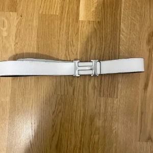 Vändbart vit bälte med H-spänne - Säljer ett vändbart bälte med ikoniskt H-spänne i silver. Ena sidan är vit och den andra svart, vilket gör det enkelt att matcha med olika outfits. Bältet är gjort i äkta skinn och har en stilren, klassisk design.