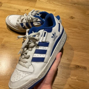 Adidas Forum Low vita och blå sneakers - Säljer ett par Adidas Forum Low sneakers i vitt och blått med klassiska tre ränder på sidan. Skorna har snörning, perforerad tå och blå detaljer på sula och foder. Perfekta för dig som gillar retrostil och streetwear. Sköna och snygga till vardags. Nästan helt nya endast använda två gånger. Box ingår.