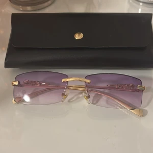 Lila rektangulära glasögon med gulddetaljer - Snygga rektangulära glasögon med lila tonade glas och guldfärgade metallbågar. Sidorna har dekorativa detaljer och näsbryggan är markerad i guld. Kommer med ett svart fodral med guldfärgad knapp.