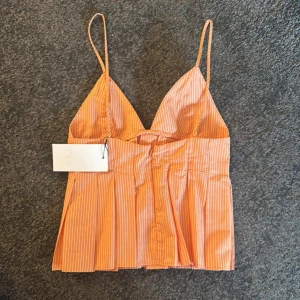 Randig orange Zara topp - Superfin orange linnetopp från Zara med tunna axelband och v-ringning. Toppen har vita smala ränder och en lätt utsvängd, plisserad nederdel som ger en snygg siluett. Perfekt till jeans eller kjol för en fräsch sommarlook.