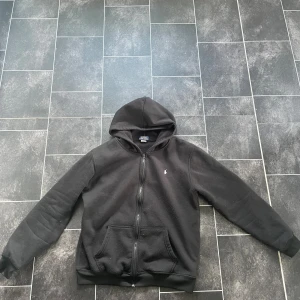 Svart hoodie från Polo Ralph Lauren - Svart hoodie från Polo Ralph Lauren med dragkedja framtill och klassisk huva. Tröjan har två fickor på magen och en liten broderad logga på bröstet. Tillverkad i mjuk polyester för en skön känsla.