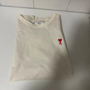 Vit t-shirt från Ami med rött hjärta - Snygg vit t-shirt från Ami med klassisk rund hals och korta ärmar. På bröstet finns Ami:s ikoniska röda hjärta med bokstaven A broderat. T-shirten är gjord i mjuk bomull och har en clean, stilren look som passar perfekt till jeans eller shorts.