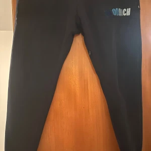 Svarta mjukisbyxor med broderad text - Svarta mjukisbyxor med broderad text i blått och vitt på ena benet. Byxorna har resår och snörning i midjan för skön passform. Perfekta för chill dagar eller när du vill vara bekväm. Tillverkade i mjukt material.Hoodrich även tröjan finns