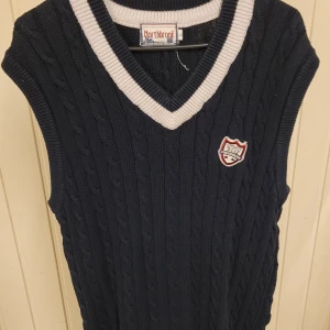 Mörkblå stickad väst från Northbrook - Snygg mörkblå stickad väst från Northbrook med klassisk kabelstickning och vit ribbad kant vid v-ringad hals. Västen har ett sportigt emblem på bröstet och är perfekt för lager-på-lager. Passar dig som gillar preppy stil och vill ha något som sticker ut.