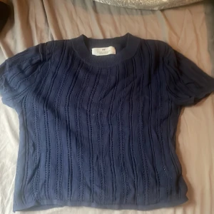Mörkblå stickad topp från H&M - Säljer en mörkblå stickad topp från H&M med korta ärmar och rund hals. Toppen har ett snyggt hålstickat mönster i vertikala ränder och är croppad i modellen. Perfekt för dig som gillar en enkel men trendig look.
