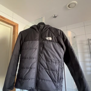 Svart pufferjacka från The North Face - Säljer en svart pufferjacka från The North Face med huva och dragkedja framtill. Jackan har en stilren look med diskret logga på bröstet och en bröstficka med dragkedja. Perfekt för kalla dagar och enkel att matcha med allt.kan vara i två olika färger som ni ser på bilderna.För mer frågor eller funderingar kontakta mig😃