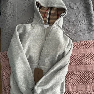 Grå hoodie från Burberry med dragkedja - Snygg grå hoodie från Burberry med klassiskt rutigt foder i huvan. Tröjan har dragkedja framtill, ribbade muddar och en mjuk bomullsblandning. Perfekt för dig som vill ha en stilren och lyxig vardagslook.