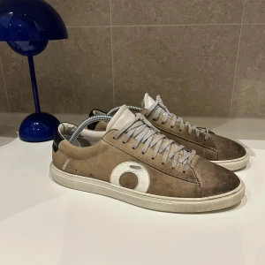 Oliver Cabell beige sneakers mocka - Snygga sneakers från Oliver Cabell i beige mocka med vit sula och grå snörning. Skorna har en vit cirkeldetalj på sidan och svart hälparti med guldfärgad logga. Perfekt för dig som gillar stilrena och unika detaljer.
