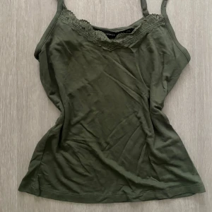 Dark sagegrön linnetopp med spetsdetalj - Snygg olivgrön linnetopp från H&M med tunna axelband och spetskant vid urringningen. Toppen är ärmlös och har en mjuk, följsam känsla i bomullsmaterial. xs/s