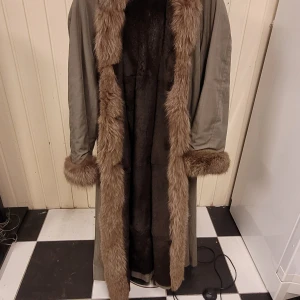 Lång pälsjacka med bruna detaljer - Säljer en lång och oversized pälsjacka i beige med brunt foder och breda pälskanter längs krage, ärmslut och öppning. Jackan har en klassisk look med mjukt material och markerade sömmar på ärmarna. Perfekt för dig som vill sticka ut med en vintagevibe.