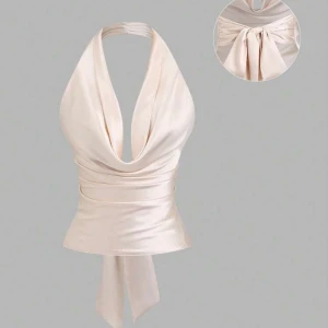 Champagnefärgad halterneck topp i satin - Snygg champagnefärgad halterneck topp i glansigt satinmaterial. Toppen har djup urringning, omlottkänsla och breda knytband i ryggen som ger en cool rosettdetalj. Perfekt för dig som vill sticka ut med en elegant och trendig look. Helt oanvänd 