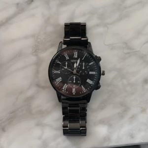 Svart klocka från Geneva med metallarmband - Stilren svart klocka från Geneva med metallarmband och romerska siffror på urtavlan. Klockan har flera små urtavlor och en klassisk rund form. Perfekt accessoar för dig som gillar en elegant och tidlös look.