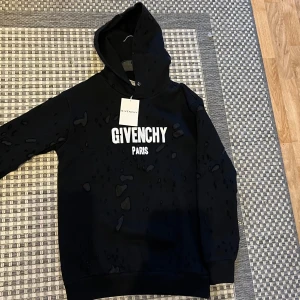 Svart hoodie från Givenchy Paris - Svart hoodie från Givenchy Paris med vit logotyptryck på bröstet. Tröjan har huva, långa ärmar och coola slitna detaljer över hela tyget som ger en edgy vibe. Tillverkad i mjuk bomull för skön komfort. Perfekt för dig som gillar streetwear och designer.