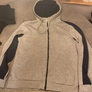 Grå hoodie från Hugo Boss med svarta detaljer - Snygg grå hoodie från Hugo Boss med svarta paneler längs ärmarna och tydligt BOSS-tryck på bröstet. Tröjan har huva med snörning, hel dragkedja och ribbade muddar. Perfekt för chill eller streetwear. Materialet är mjuk bomull och polyester.
