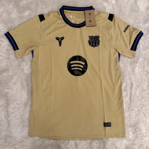Beige FC Barcelona fotbollströja Nike - Snygg beige FC Barcelona matchtröja från Nike med svarta och blå detaljer. Kortärmad modell med klubbmärke, Spotify-logga och UNHCR-tryck. Rund hals med blå och svart kant. Perfekt för dig som vill ha en unik Barca-tröja med modern design.