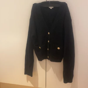 Svart kofta med guldknappar H&M - Svart stickad kofta från H&M i storlek S. Koftan har snygga guldknappar framtill och två fickor. Den är ribbstickad och har en klassisk v-ringning samt långa ärmar. Perfekt att slänga på sig när det är kyligt ute.
