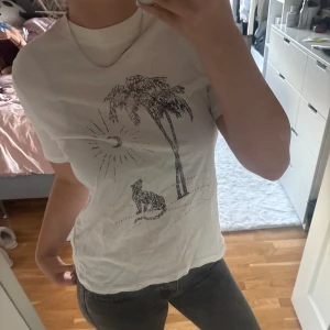 Vit t-shirt med palm- och leopardtryck - Vit t-shirt med korta ärmar med ett coolt svart tryck framtill. T-shirten har en klassisk passform och funkar bra till både jeans och kjol. 