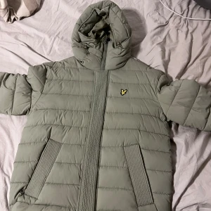 Ljusgrön pufferjacka från Lyle & Scott - Snygg ljusgrön pufferjacka från Lyle & Scott med huva och dragkedja framtill. Jackan har quiltad design, två stora fickor och den klassiska gula loggan på bröstet. Tillverkad i nylonblandning och perfekt för kyliga dagar.