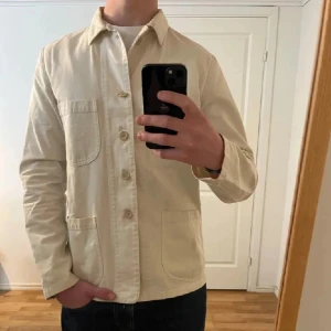Beige overshirt från Alberto Aspesi - Stilren beige overshirt från Alberto Aspesi i bomull. Jackan har klassisk krage, tre stora utanpåfickor och knäppning framtill med fyra knappar. Perfekt för lager-på-lager och enkel att matcha med jeans eller chinos. Saknar halva knappen på en av de översta o lätt fläck vid nacken, annars bra skick!!