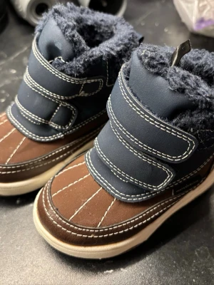 Blåbruna vinterboots från H&M, stl 20-21 - Säljer ett par blå och bruna vinterboots från H&M med mysigt foder och två kardborreband för enkel påtagning. Skorna har vit sula, dekorativa sömmar och är perfekta för kalla dagar. Utsidan är i syntetmaterial och insidan är fodrad för extra värme.