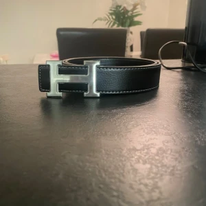 Svart h-ermes bälte - Snyggt svart skärp med ett stort, silvrigt H-format spänne. Skärpet har vita sömmar längs kanterna och är gjort i ett material som liknar skinn. Perfekt accessoar för att lyfta din outfit.