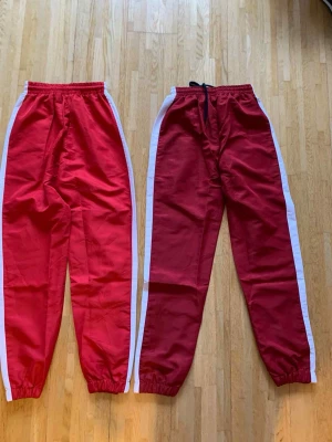 Röda Lacoste träningsbyxor med vita ränder - Säljer två par röda träningsbyxor från Lacoste med vita ränder längs sidorna och elastisk midja. Ena paret har snörning i midjan. Klassisk Lacoste-logga på låret. Perfekta för en sportig look och riktigt sköna att bära.