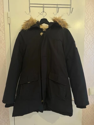 Svea vinter parkas Miss Smith Black med fuskpäls – elegant, varm & snygg - Produktbeskrivning 🧥Snygg, varm och stilren svart vinterjacka från Svea. 🧥Har beige foder och huva med fuskpäls. 🧥Köpt från Junkyard. 🧥Nypris är 4599kr. 🧥Jackan är slutsåld i flera ställen. 🧥Bröstmått: ca 50cm 🧥Längd: ca 75cm 🧥Jackan har stora fickor framtill, dragkedja och knappar samt en patch med amerikansk flagga på ärmen. 🧥I ganska fin skick — inga synliga defekter.  🧥Jackan är en casual men ändå fashionabel och kombinerar värme samt komfort med en tidlös look. 🧥Perfekt för vardagsbruk.