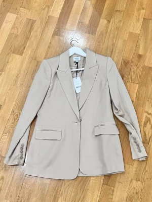 Beige kavaj från NA-KD Reborn, strl 36 - Snygg beige kavaj från NA-KD Reborn i storlek 36. Klassisk modell med breda slag, knäppning framtill och fickor med lock. Kavajen har fyra knappar vid ärmsluten och är tillverkad i ett mjukt material som ger en clean och stilren look.