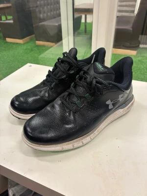 Svarta sneakers från Under Armour - Säljer ett par svarta sneakers från Under Armour med vit sula och gröna detaljer på snörningen. Skorna har en sportig look med rund tå och är tillverkade i skinn och syntet. Perfekta för dig som gillar stilrena och bekväma skor till vardags.