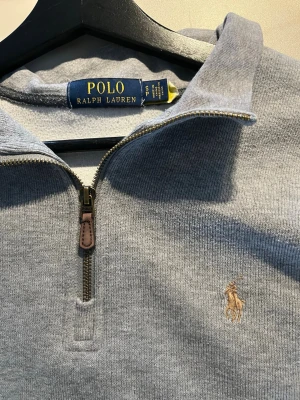 Grå half zip tröja från Polo Ralph Lauren - Snygg grå tröja från Polo Ralph Lauren med half zip och klassisk broderad logga på bröstet. Tröjan har lång ärm och en stilren krage. Perfekt för lager-på-lager och passar till jeans eller chinos.