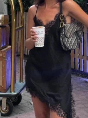 Svart slipklänning med spetsdetaljer S - Säljer en svart kort slipklänning med tunna axelband och vacker spets längs urringning och nederkant. Klänningen har asymmetrisk fåll och en mjuk, följsam känsla. Perfekt för dig som gillar en chic och enkel look med lite extra edge.