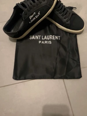 Svarta Saint Laurent sneakers - Svarta sneakers från Saint Laurent med vit logga på sidan och vit sula. Skorna har snörning och är tillverkade i skinn och textil. Medföljer original dustbag med Saint Laurent Paris-tryck. Perfekta för dig som gillar stilrena och exklusiva sneakers.
