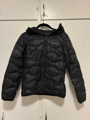 Svart pufferjacka Peak Performance - Säljer en svart pufferjacka från Peak Performance med huva och dragkedja framtill. Jackan har quiltad design och är lätt vadderad, perfekt för kyliga dagar. Klassisk passform och diskret logga på bröstet. Grym jacka för dig som vill ha både stil och funktion.