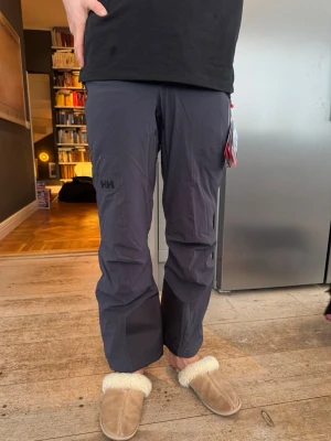 Mörkgrå/blå skidbyxor från Helly Hansen - Säljer ett par mörkblå skidbyxor från Helly Hansen. Helt nya och aldrig använda. Funktionella skidbyxor av hög kvalitet, designade för värme, komfort och bra prestanda i backen.  Observera: byxorna är professionellt förkortade för modellen som är 194 cm lång. Detta gör dem passande för någon som är något kortare, beroende på önskad passform och pjäxor. Perfekta inför skidsäsongen som börjar nu. 
