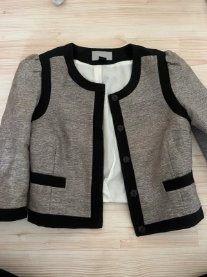 Grå och svart kavaj med knappar - Snygg grå kavaj med svarta detaljer och dekorativa knappar framtill. Jackan har rundad halsringning, långa ärmar och två fickor med svarta kanter. Materialet känns mjukt och har en lätt struktur. Perfekt för dig som vill ha en stilren och modern look.