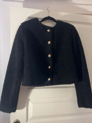 Svart kofta från H&M med guldknappar - Säljer en svart, fluffig kofta från H&M i storlek S. Koftan har långa ärmar, rund halsringning och snygga guldknappar framtill. Materialet är mjukt och ger en mysig känsla, perfekt för kyligare dagar.