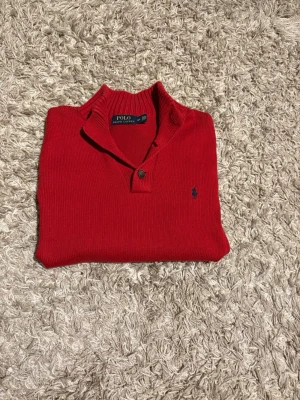  stickad tröja från Polo Ralph Lauren - Snygg röd stickad tröja från Polo Ralph Lauren med klassisk krage och knappdetalj vid halsen. Tröjan har en broderad logga på bröstet och är perfekt för dig som gillar stilrena och tidlösa plagg.