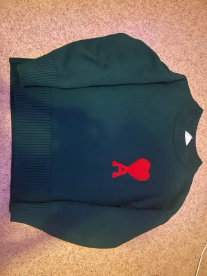 Mörkgrön stickad tröja från Ami - Snygg mörkgrön stickad tröja från Ami Alexandre Mattiussi med ett stort rött hjärta och bokstaven A broderat på bröstet. Tröjan har ribbade muddar vid ärmslut och nederkant samt rund hals. Perfekt för dig som gillar stilrena detaljer och vill sticka ut.