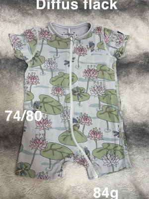 Kortärmad blommig pyjamas 74/80 - Supersöt kortärmad pyjamas med dragkedja framtill och ett gulligt mönster av näckrosor och trollsländor i grönt, rosa och vitt. Perfekt för varma dagar och enkel att ta på tack vare dragkedjan.🌸 Diffus fläck. Storlek 74/80