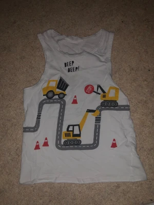Barn tröja - Säljer en vit linnetopp från H&M i storlek 134/140. Toppen har tryck med gula byggfordon, vägar, koner och texten 'BEEP BEEP!' framtill. Materialet är mjuk bomull och modellen är ärmlös med rund hals. Perfekt för dig som gillar coola motiv.