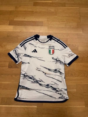 Italien Adidas fotbollströja 13-14 år - Säljer en snygg Italien fotbollströja från Adidas i vitt med marinblå marmormönster och detaljer. Tröjan har klassiska Adidas-ränder på axlarna, Italiens landslagsemblem på bröstet och är tillverkad i lätt polyester. Perfekt för dig som älskar fotboll och vill stötta Italien!