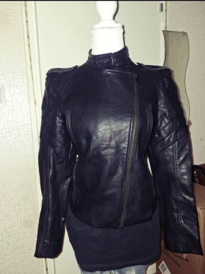 Svart Zara skinnjacka med dragkedja - Säljer en svart skinnjacka med coola detaljer och snedställd dragkedja. Jackan har markerade axlar, quiltade partier på ärmarna och ståkrage med knapp. Passformen är normal och jackan har flera dragkedjor framtill för en edgy look.