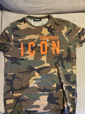 DSQUARED2 camo t-shirt ICON 12Y - Säljer en cool t-shirt från DSQUARED2 i storlek 12Y med klassiskt camouflage-mönster i grönt, brunt och beige. Framsidan har orange tryck med texten 'ICON'. T-shirten är kortärmad och har rund hals. Perfekt för dig som gillar streetwear och statement-plagg. Ej använd på år. 